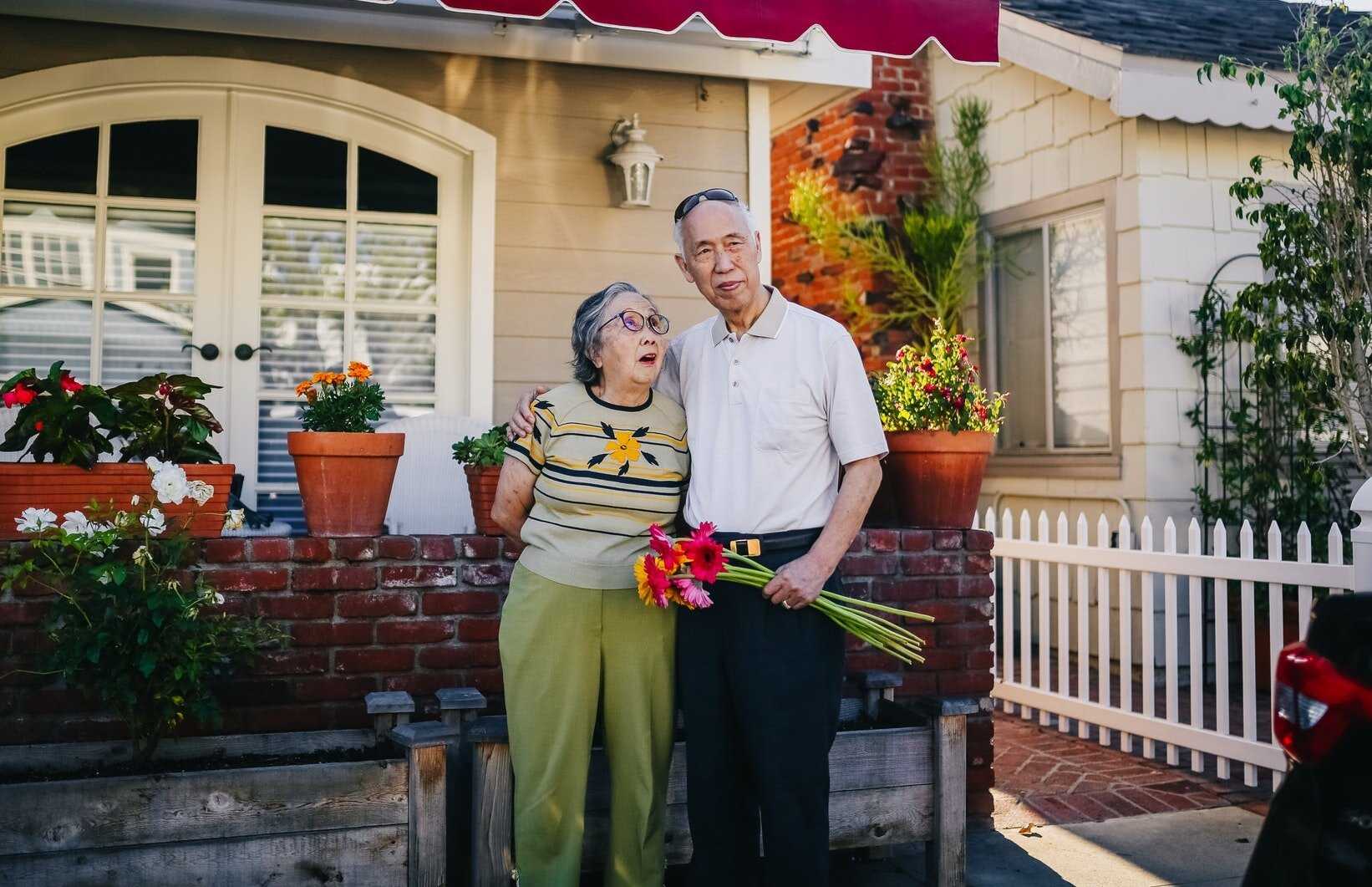 Two elderly adults standing in front of their home and holding flowers that they have grown.