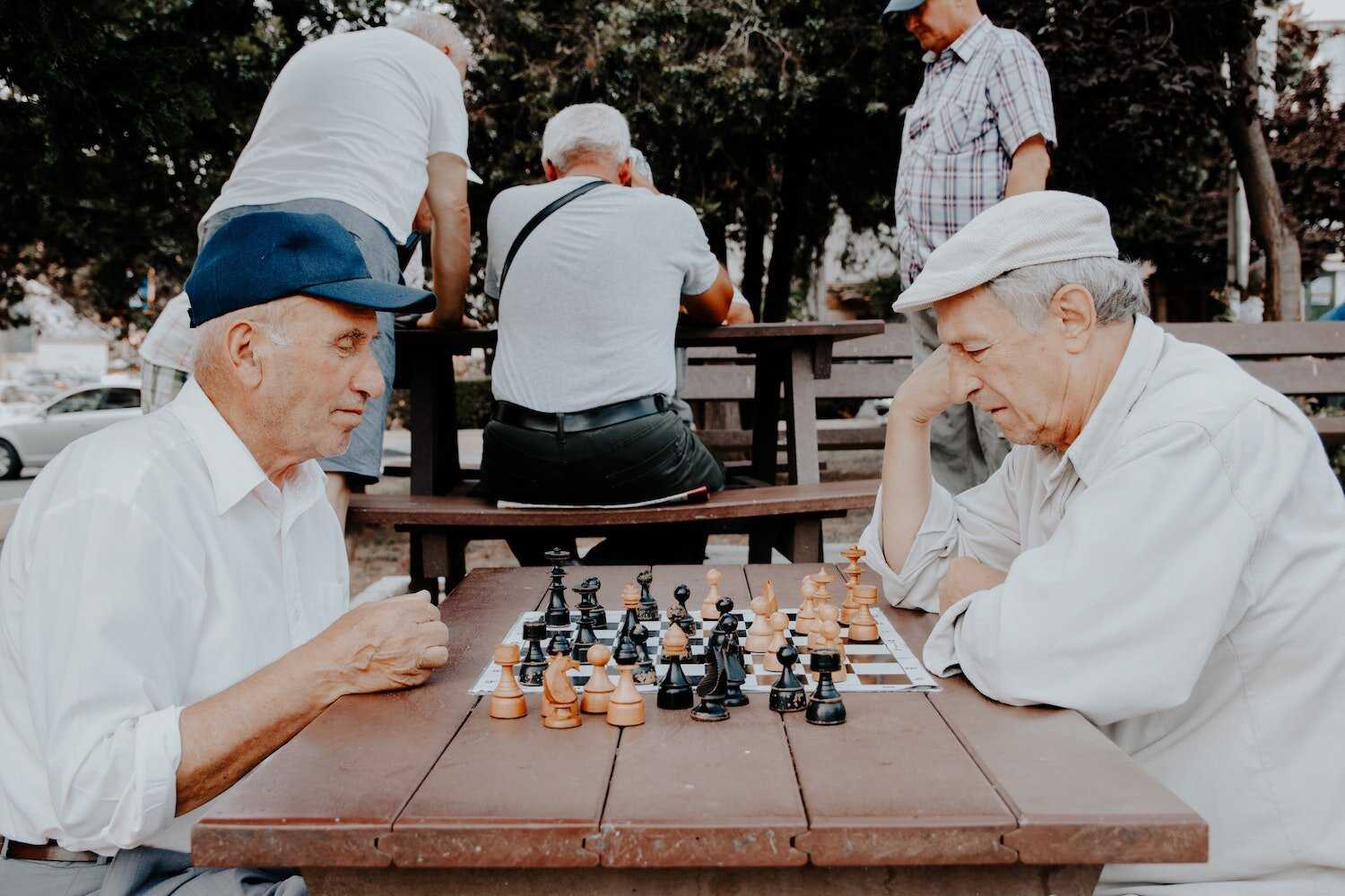 Two elderly men in the park playing chess together while others are playing in the background.